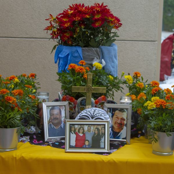Día de los Muertos altar decorated with vibrant marigolds and red flowers, framed photographs of loved ones, a decorative skull, candles, and a wooden cross, all arranged on a yellow cloth-covered table.