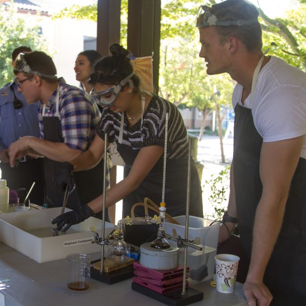 Students wearing safety goggles and aprons conducting a chemistry demonstration outdoors, using glass beakers, tubing, and lab equipment on a table