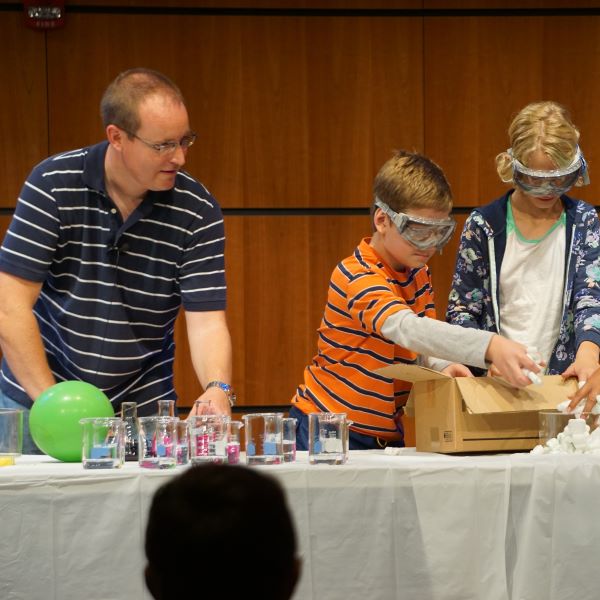 A man and two children participate in a science experiment, wearing safety goggles and working with beakers and a box of materials on a table.